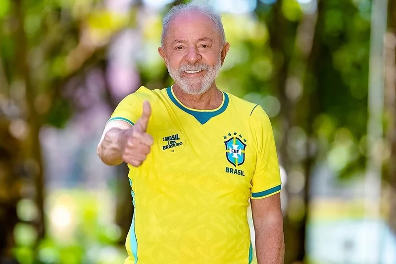 Smiling older man in a Brazil national team jersey giving a thumbs up outdoors.