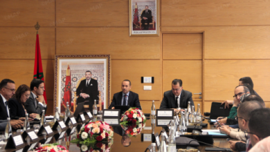 Formal boardroom meeting with a man speaking at the head of a long table, Moroccan flag and portraits on a wood-paneled wall.