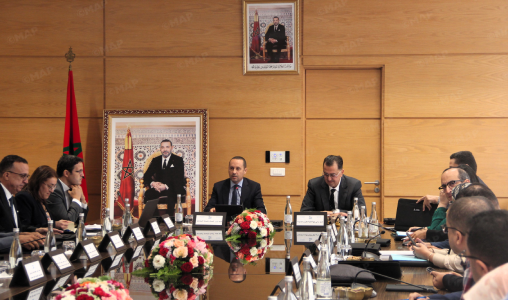 map99991 | MCG24 | موقع إلكتروني مغربي شامل متجدد على مدار الساعة Formal boardroom meeting with a man speaking at the head of a long table, Moroccan flag and portraits on a wood-paneled wall.
