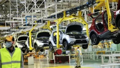 Car manufacturing assembly line with vehicles on an overhead conveyor and workers in high-visibility vests inspecting them in a factory.
