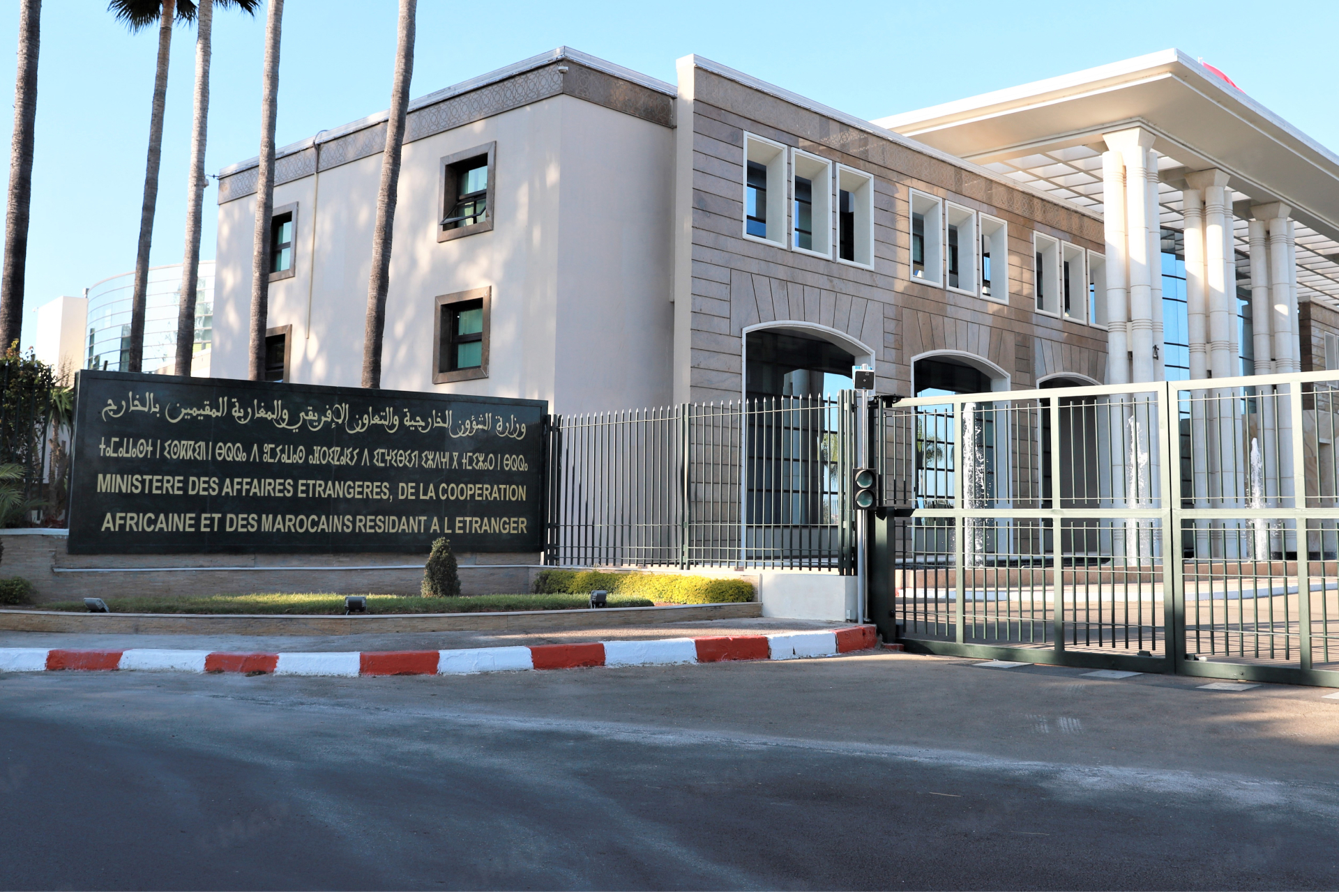 Front view of a modern government building behind a tall metal fence with a large sign in Arabic and French listing the Ministry of Foreign Affairs and Cooperation for Moroccans abroad.