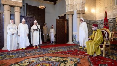 Moroccan king in yellow robes sits on a throne in a richly decorated hall, with officials standing and guards nearby.