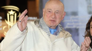 morin-a-marrakech-1780136438 | MCG24 | موقع إلكتروني مغربي شامل متجدد على مدار الساعة Elderly man in a cream coat and striped scarf waves at the camera indoors.