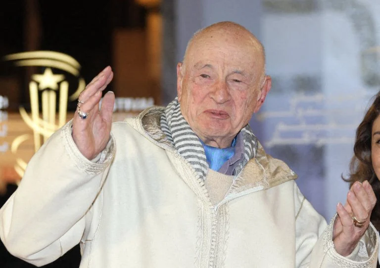 Elderly man in a cream coat and striped scarf waves at the camera indoors.
