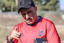 Man in a red sports jersey points at the camera outdoors, wearing a black cap and holding a tablet.