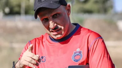 جعواني | MCG24 | موقع إلكتروني مغربي شامل متجدد على مدار الساعة Man in a red sports jersey points at the camera outdoors, wearing a black cap and holding a tablet.