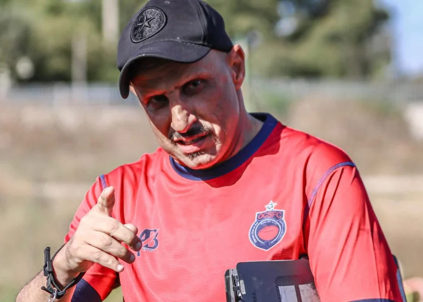 Man in a red sports jersey points at the camera outdoors, wearing a black cap and holding a tablet.