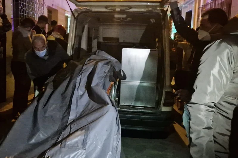 People wearing masks load a body covered by a gray tarp into a van at night, while bystanders watch nearby.
