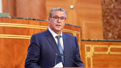 Man in a dark blue suit and glasses delivering a speech at a wooden podium with microphones in a formal setting