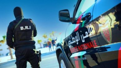 polic-535x356 | MCG24 | موقع إلكتروني مغربي شامل متجدد على مدار الساعة Tactical police officer in a vest labeled B.R.I. stands beside a black police SUV with red-green stripes and Arabic text.