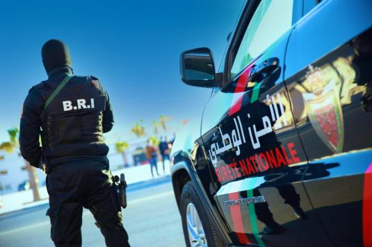 polic-535x356 | MCG24 | موقع إلكتروني مغربي شامل متجدد على مدار الساعة Tactical police officer in a vest labeled B.R.I. stands beside a black police SUV with red-green stripes and Arabic text.