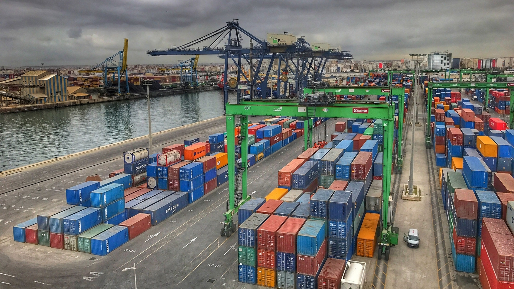 Aerial view of a busy container port with colorful shipping containers and large green gantry cranes along a waterway under a cloudy sky.