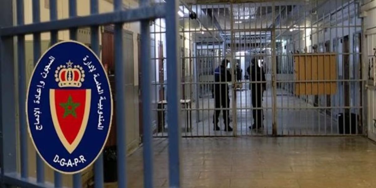 prison-1 2 | MCG24 | موقع إلكتروني مغربي شامل متجدد على مدار الساعة Prison corridor behind a metal-barred gate with a blue DGAPR emblem in the foreground and two people standing in the hall behind the gate.