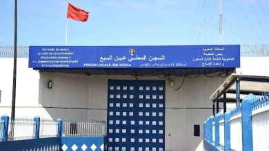 prison-AIN-SBAA | MCG24 | موقع إلكتروني مغربي شامل متجدد على مدار الساعة Front view of Ain Sebaa local prison in Morocco: blue sign in Arabic and French above a solid blue gate, red national flag flying nearby, surrounded by a spiked fence and blue railings.