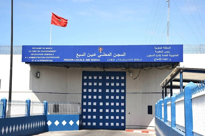 prison-AIN-SBAA | MCG24 | موقع إلكتروني مغربي شامل متجدد على مدار الساعة Front view of Ain Sebaa local prison in Morocco: blue sign in Arabic and French above a solid blue gate, red national flag flying nearby, surrounded by a spiked fence and blue railings.