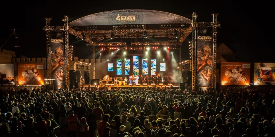 Outdoor nighttime concert with a large crowd facing a well-lit stage and multiple vertical video screens.