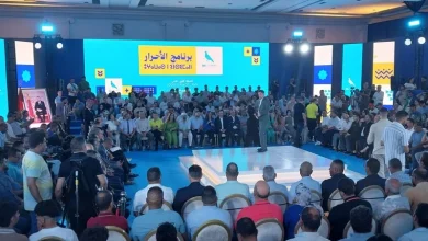 rni-fes | MCG24 | موقع إلكتروني مغربي شامل متجدد على مدار الساعة Speaker on a raised white platform speaks to a large audience seated in a circular arrangement in a conference hall; blue stage backdrop with screens displaying Arabic text.