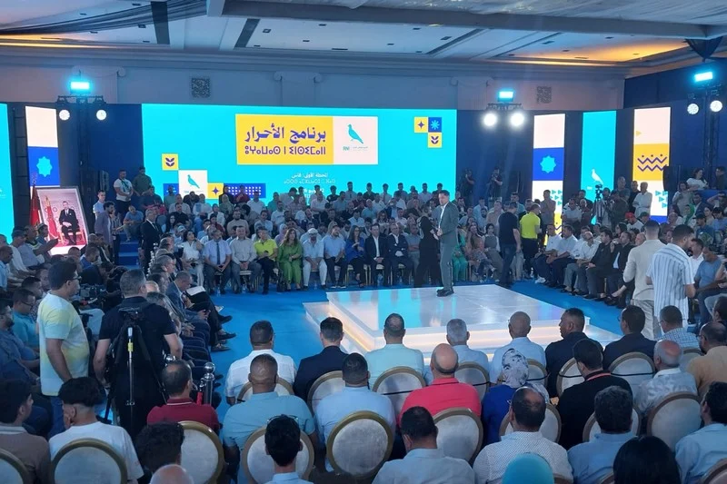 rni-fes | MCG24 | موقع إلكتروني مغربي شامل متجدد على مدار الساعة Speaker on a raised white platform speaks to a large audience seated in a circular arrangement in a conference hall; blue stage backdrop with screens displaying Arabic text.