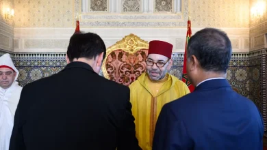Man in a yellow Moroccan djellaba and red fez converses with two men in suits before a gilded throne in an ornate room with tilework and patterned wallpaper.