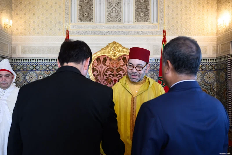 Man in a yellow Moroccan djellaba and red fez converses with two men in suits before a gilded throne in an ornate room with tilework and patterned wallpaper.