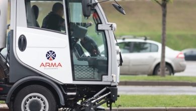 s1 | MCG24 | موقع إلكتروني مغربي شامل متجدد على مدار الساعة White street-sweeping truck in operation, brushing the road surface with a large undercarriage broom, driver visible inside the cab (ARMA Environmental logo on the side).