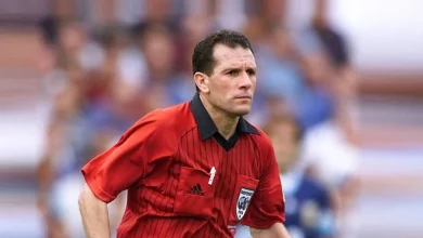 بلقولة | MCG24 | موقع إلكتروني مغربي شامل متجدد على مدار الساعة Soccer referee in a red striped jersey runs on the field, with a blurred crowd in the background.