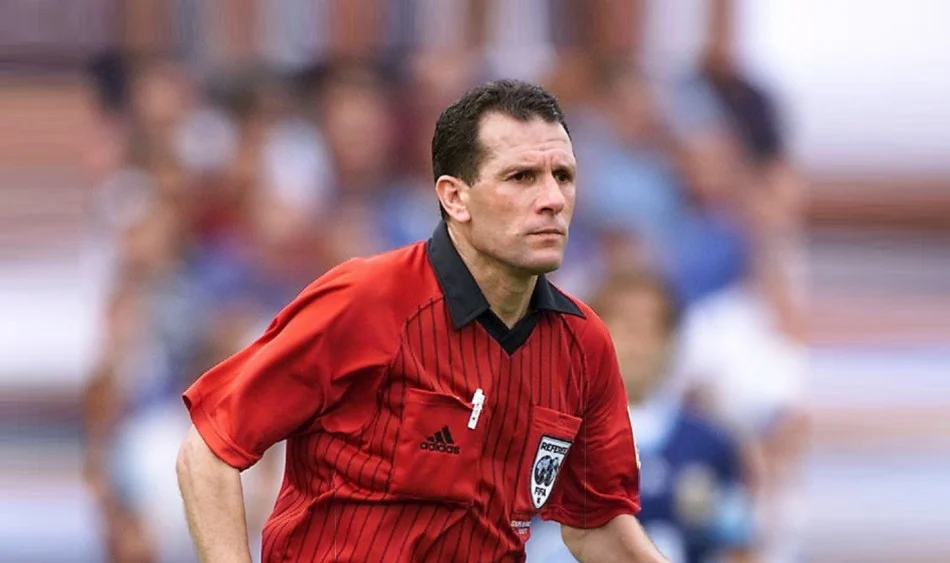 بلقولة | MCG24 | موقع إلكتروني مغربي شامل متجدد على مدار الساعة Soccer referee in a red striped jersey runs on the field, with a blurred crowd in the background.