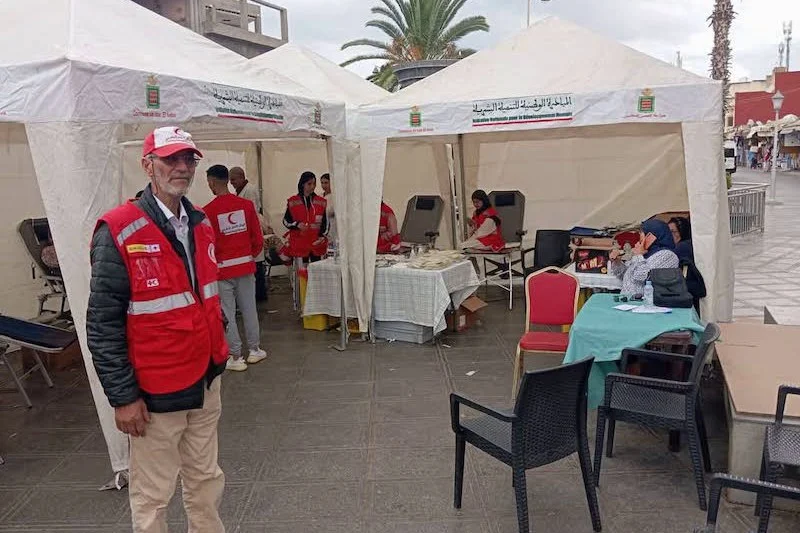 sang-4 | MCG24 | موقع إلكتروني مغربي شامل متجدد على مدار الساعة Outdoor medical aid tents with volunteers in red vests; a man wearing a red cap stands in foreground while others assist at tables under white canopies