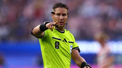Football referee in a bright neon yellow jersey points toward the play, wearing a headset and a FIFA badge on the chest.