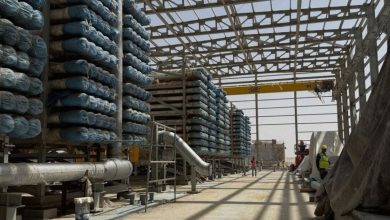 444777 | MCG24 | موقع إلكتروني مغربي شامل متجدد على مدار الساعة Industrial construction site with rows of stacked blue-wrapped pipes and a yellow overhead crane in a partially built steel framework.