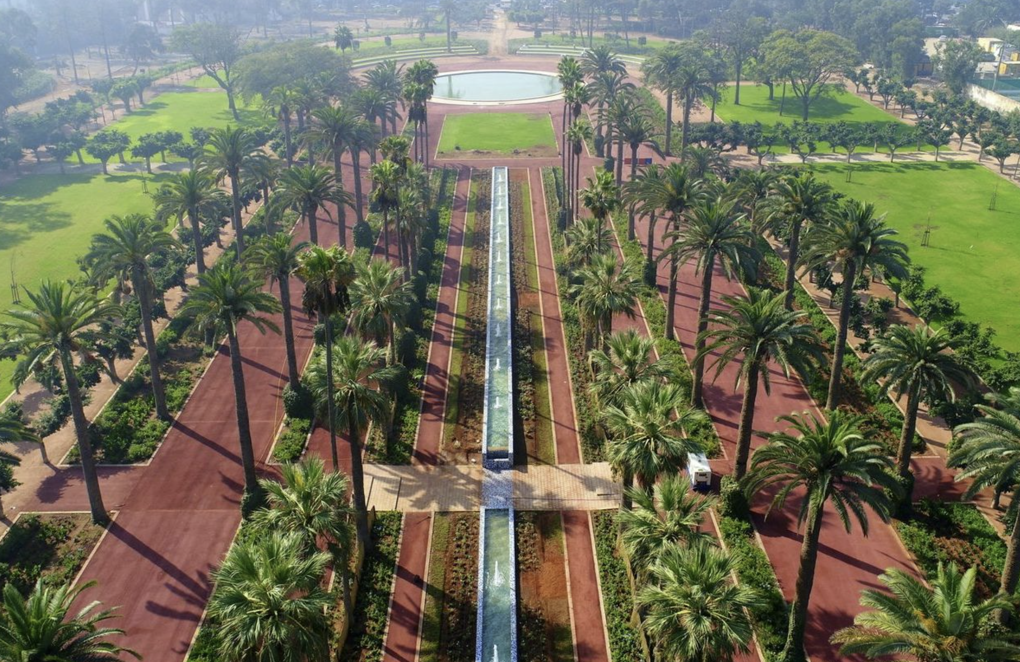 screenshot-2026-06-18-123636 | MCG24 | موقع إلكتروني مغربي شامل متجدد على مدار الساعة Aerial view of a formal park with red brick paths, tall palm trees, and a central long fountain leading to a circular pond.