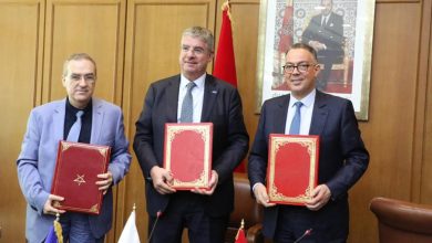 signature-post-eath768 | MCG24 | موقع إلكتروني مغربي شامل متجدد على مدار الساعة Three men in suits stand in a wood-paneled room, each holding a red ornate folder during a formal signing ceremony with flags in the background.