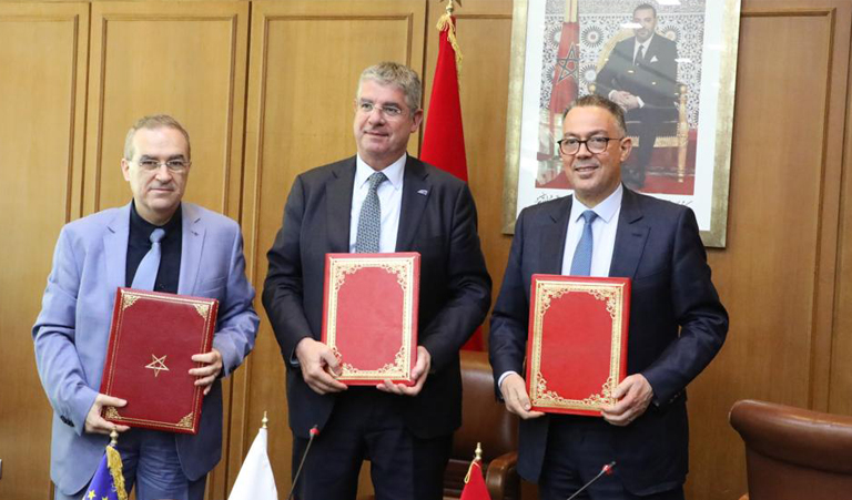 signature-post-eath768 | MCG24 | موقع إلكتروني مغربي شامل متجدد على مدار الساعة Three men in suits stand in a wood-paneled room, each holding a red ornate folder during a formal signing ceremony with flags in the background.