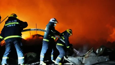 Firefighters in protective gear battle a large blaze at night, spraying water on smoldering rubble against an orange sky