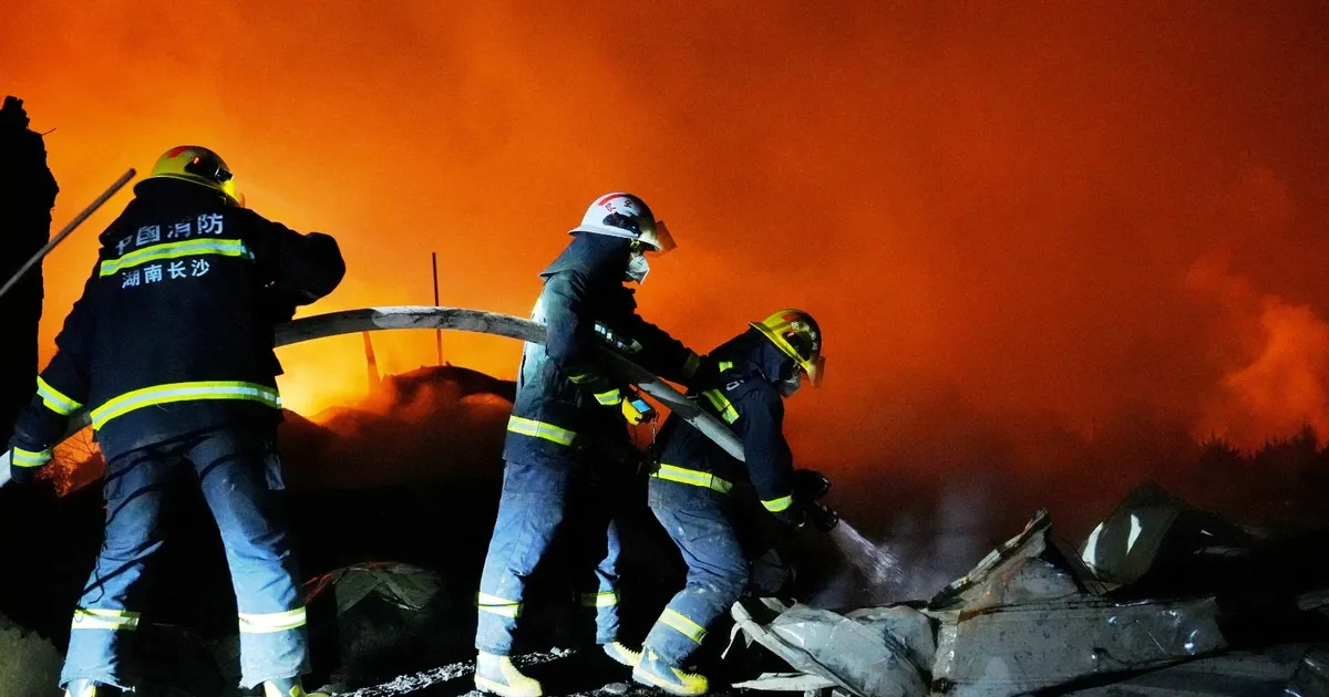 Firefighters in protective gear battle a large blaze at night, spraying water on smoldering rubble against an orange sky
