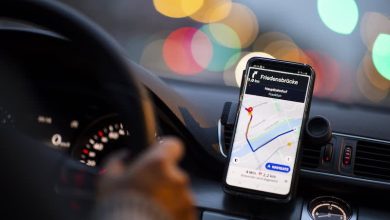 transport-application | MCG24 | موقع إلكتروني مغربي شامل متجدد على مدار الساعة Phone mounted on a car dashboard displaying a navigation app with a route on a map in focus, city lights blurred in the background.