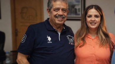 unnamed 8 | MCG24 | موقع إلكتروني مغربي شامل متجدد على مدار الساعة Smiling man in a navy polo stands next to a woman in an orange shirt inside an indoor room with framed photos on the wall.