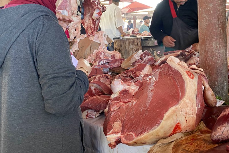 viande-2 | MCG24 | موقع إلكتروني مغربي شامل متجدد على مدار الساعة Meat market stall with large cuts of beef on a table; a customer in a gray hoodie selects meat while others shop in the background.