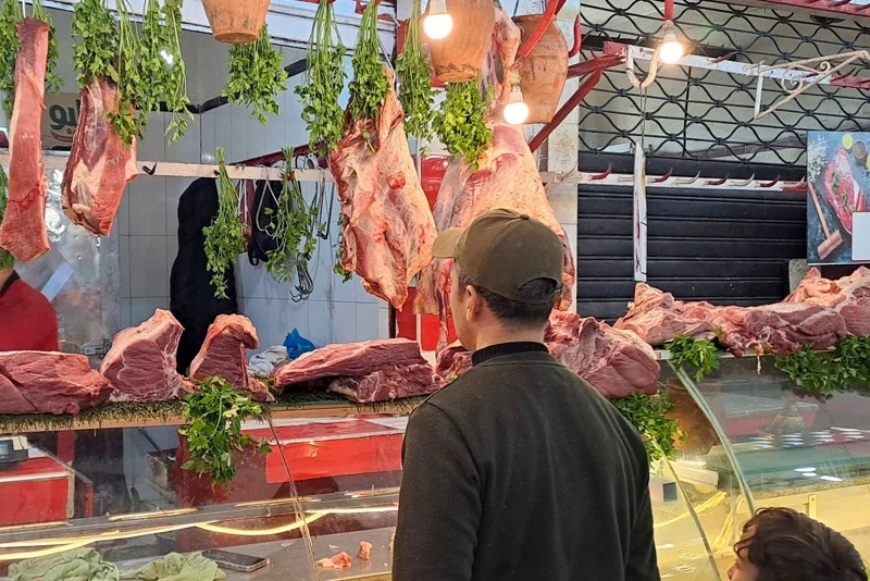 viande | MCG24 | موقع إلكتروني مغربي شامل متجدد على مدار الساعة Butcher shop scene with large meat cuts hanging from hooks above a glass display case, herbs dangling nearby, and a man in a cap watching the counter.
