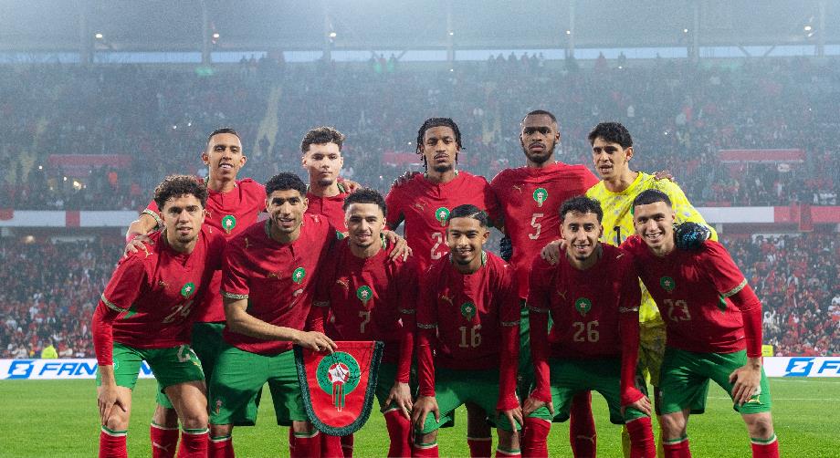 Morocco national football team posing for a group photo on the field with a shield banner in front of a crowded stadium