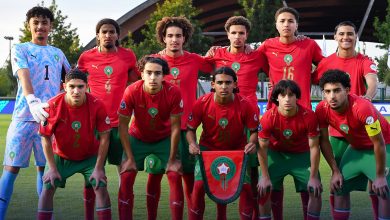 vis_0523202611544614 | MCG24 | موقع إلكتروني مغربي شامل متجدد على مدار الساعة Group of eleven soccer players posing for a team photo on a grassy field; red jerseys with green shorts, goalie in blue jersey on far left.