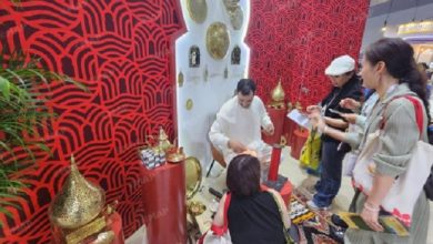 vis_0529202611423654 | MCG24 | موقع إلكتروني مغربي شامل متجدد على مدار الساعة Man in white arranges gold offerings on a small altar as three customers observe, in a shop with bold red patterned walls and ornate decorative items.
