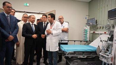 Group of suited officials listening to a doctor in a white coat in a hospital ICU beside an empty bed and monitors.