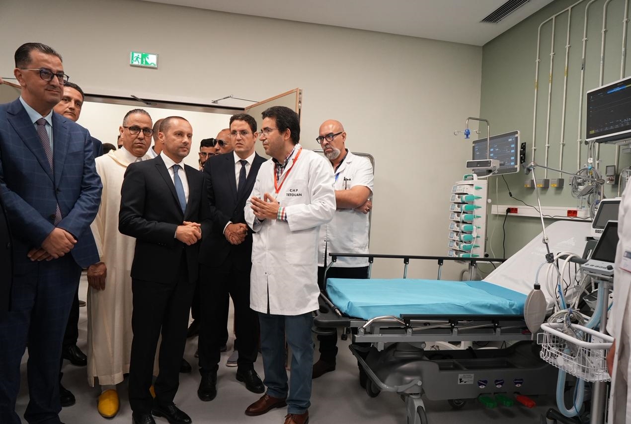 Group of suited officials listening to a doctor in a white coat in a hospital ICU beside an empty bed and monitors.