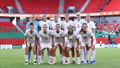 Group of female soccer players in white jerseys with green trim posing for a team photo on a soccer field inside a stadium with red and green seating; goalkeeper in blue stands at left back.