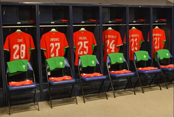 vis_0607202618264718 | MCG24 | موقع إلكتروني مغربي شامل متجدد على مدار الساعة Row of navy lockers with red football jerseys and numbers 28, 27, 25, 24, 23; green duffel bags on chairs in front of each locker.
