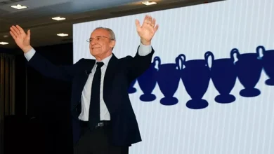 848 | MCG24 | موقع إلكتروني مغربي شامل متجدد على مدار الساعة Older man in a dark suit with arms raised in celebration on a stage, backdrop showing a row of blue trophy silhouettes.