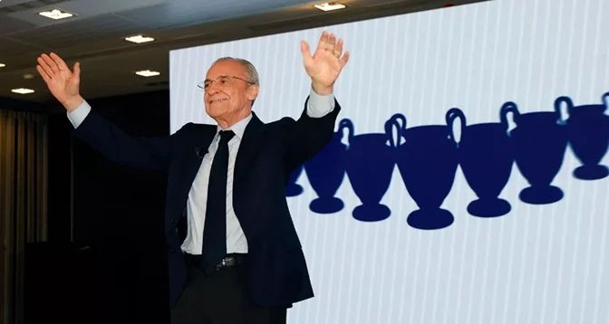 848 | MCG24 | موقع إلكتروني مغربي شامل متجدد على مدار الساعة Older man in a dark suit with arms raised in celebration on a stage, backdrop showing a row of blue trophy silhouettes.