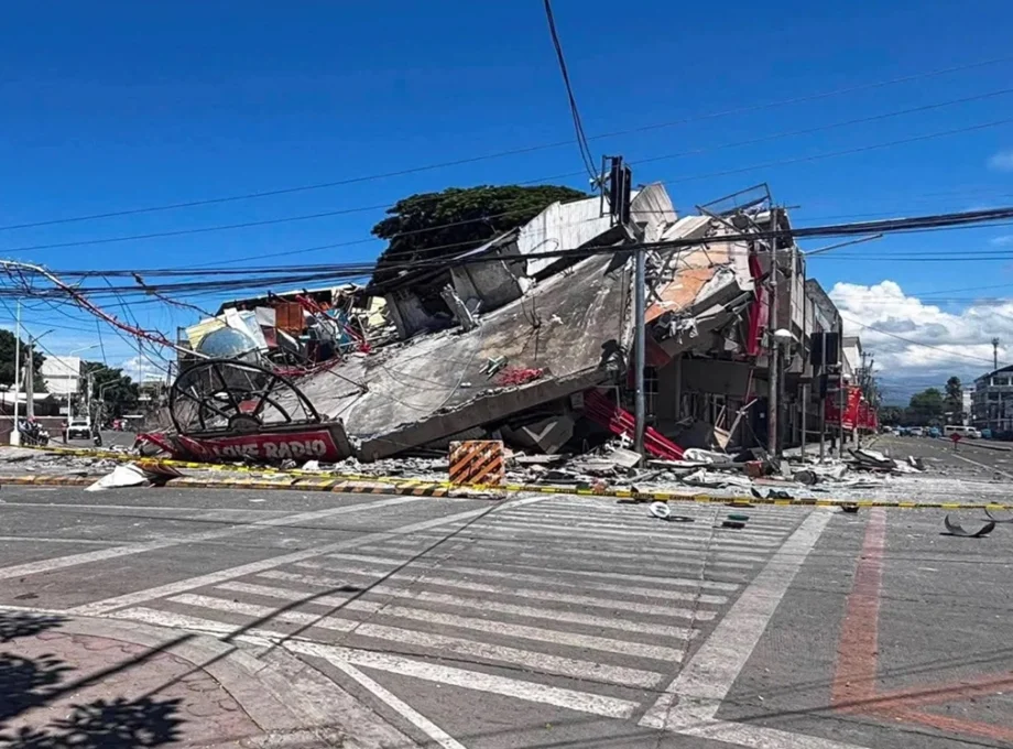 Collapsed building and rubble blocking a city street with yellow caution tape and overhead power lines.",