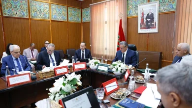 vis_0609202619525930 | MCG24 | موقع إلكتروني مغربي شامل متجدد على مدار الساعة Formal meeting around a wooden conference table with officials, Moroccan flag, and a framed portrait in an ornate wood-paneled room.